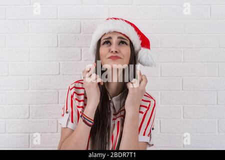 Nahaufnahme Porträt der schönen jungen Frau trägt roten weihnachtsmann Hut, die Finger zu drücken, Augen zu schließen, beten für ein Wunder, isoliert auf weiß Stockfoto