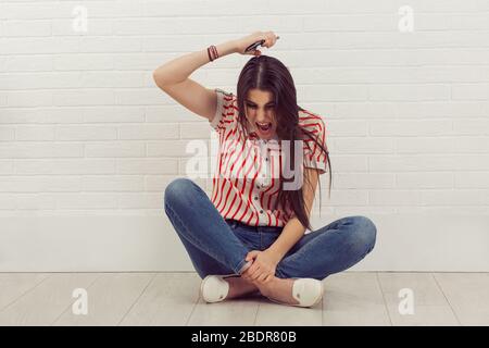 Porträt eine böse junge Frau schreit auf Handy, um ihr Telefon auf dem Boden isoliert über weißen Ziegelsteinen Hintergrund wegwerfen. Negative Emotionen Stockfoto