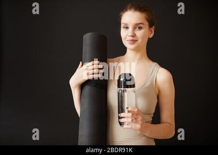 Porträt einer jungen gesunden Frau, die Übungsmatte und eine Flasche Wasser in den Händen hält und die Kamera auf schwarzem Hintergrund betrachtet Stockfoto