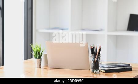 Computer-Laptop, Bleistifthalter, Topfpflanze, Notebook und Kaffeetasse zusammen auf modernen Holztisch mit Büro-Bücherregal als Hintergrund. Stockfoto