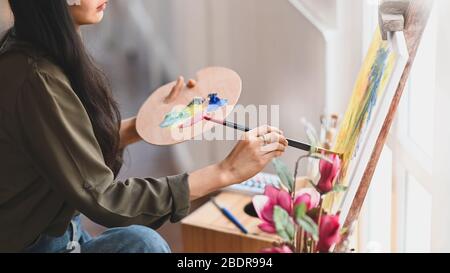 Zugeschnittenes Bild eines jungen Künstlermädchens, das einen Pinsel hält und Ölfarben auf Leinwand zeichnet, während es im Studio für moderne Kunst sitzt. Konzept des Creat Stockfoto
