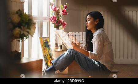 Foto von jungen Künstlermädchen, das einen Pinsel hält und Ölfarben auf Leinwand zeichnet, während sie im modernen Kunststudio sitzt. Konzept der kreativen woma Stockfoto