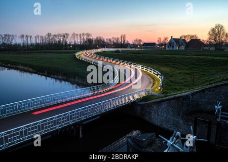Geschwungene asphalanische Landstraße in holländischer Landschaft mit weißem Zaun, der zu einem alten Bauernhaus in Holland führt. Stockfoto