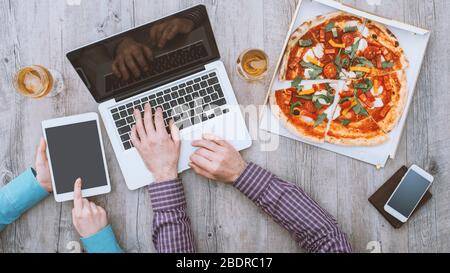 Büroangestellte, die eine Mittagspause im Büro haben, essen Pizza, verwenden einen Laptop und ein digitales Tablet, flaches Lay Stockfoto