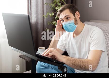 Junge kaukasische düstere Mann in Stress arbeitet aus dem Home Office tragen Schutzmaske mit Laptop und Internet. Coronavirus Covid 19 Quarantäne Stockfoto