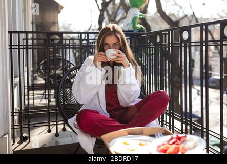 Süße zarte Dame am Morgen auf dem Balkon sitzen und aromatischen heißen Tee trinken. Tragen weißen Bademantel und burgunderroten Pyjama. Leckeres Frühstück Stockfoto