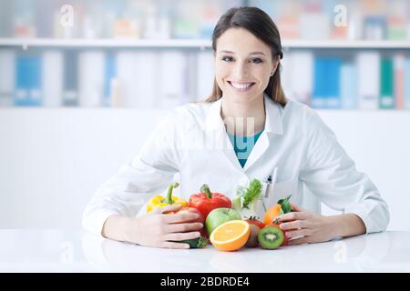 Lächelnde Ernährungsberaterin in ihrem Büro hält sie gesundes Gemüse und Obst, Gesundheitspflege und Diätkonzept Stockfoto