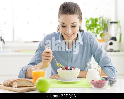 Lächelnde, glückliche Frau, die zu Hause ein köstliches gesundes Frühstück hat, isst Müsli und Cornflakes, Wtih Obst und Joghurt und sitzt am Küchentisch Stockfoto