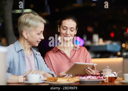 Junge Frau mit digitalen Tablet zeigt etwas zu reifen Frau, während sie am Tisch mit Tee im Café sitzen Stockfoto