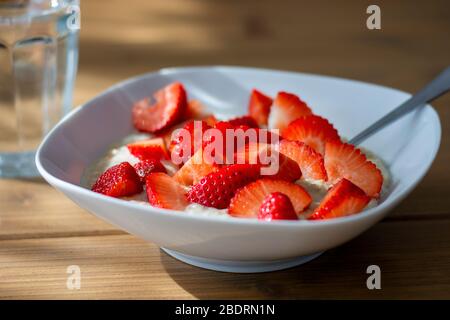 Hausgemachte Haferbrei Haferbrei Schüssel mit Erdbeeren in Scheiben geschnitten und ein Glas Wasser auf dem Tisch in der Morgensonne. Gesunde Mahlzeit. Haferbrei Stockfoto