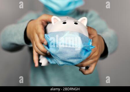 Ein kleiner Junge, der ein Sparschwein in der Gesichtsmaske hält. Symbol für schlechtes Haushaltsbudget. Persönliche Budgetierung während der Coronavirus Pandemie. COVID-19 Verletzten Famil Stockfoto