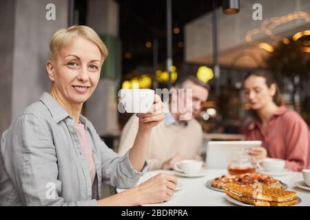 Porträt einer reifen Frau, die eine Tasse Tee hält und bei der Kamera lächelt, während sie mit ihren Freunden im Café zu Mittag essen Stockfoto