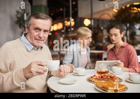 Porträt des reifen Mannes, der Tee trinkt und die Kamera anschaut, während er mit seiner Familie im Café zu Mittag essen Stockfoto