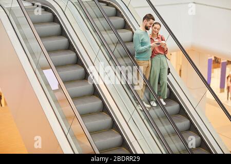 Junges Paar mit Kaffeegetränken, das auf der Rolltreppe im Einkaufszentrum steht Stockfoto