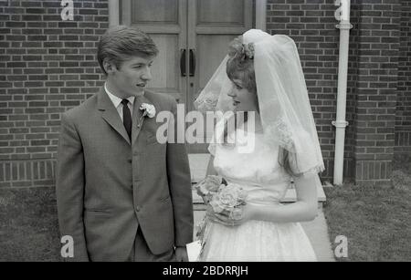 1960er Jahre, historische, "Teenager" Braut in ihrem Hochzeitskleid mit Brautschleier und Bräutigam in einem drei-Knopf-Anzug der Zeit, zusammen vor dem Eingang zu einem modernen Kirchengebäude, England, Großbritannien. Stockfoto