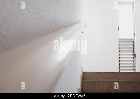Weißes Licht aus einem Fenster erhellt die weißen, minimalistischen Wände einer Gebäudetreppe. Stockfoto