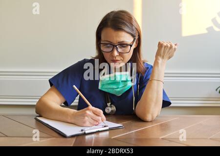 Ärztin in blauer Uniform, medizinische Maske am Tisch sitzend, Schrift auf Klemmbrett Stockfoto