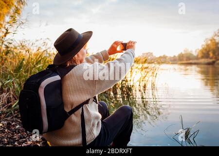 Tourist mit Rucksack, Fotos mit dem Smartphone des Flusses bei Sonnenuntergang. Frau reist und bewundert die Frühlingsnatur Stockfoto