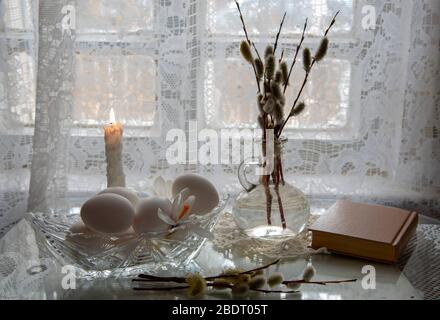 Weiße Eier in einer Kristallvase, Weidenzweige in einer Flasche und ein altes Buch auf hellem Hintergrund. Stockfoto