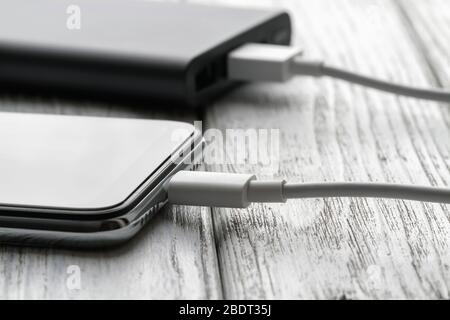 Das Smartphone lädt von einer externen Powerbank mit Kabel auf einem grauen Holzhintergrund. Nahaufnahme, selektiver Fokus Stockfoto