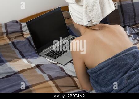 Junge schöne Frau liegt nach dem Bad mit einem gewickelten Handtuch auf dem Kopf und mit einem Laptop in einem Bett. Zeit während der Quarantäne verbringen. Bleiben Sie zu Hause Stockfoto