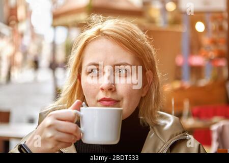 Junge schöne weiße kaukasische Frau mit blonden Haaren sitzt im Café draußen mit einer kleinen Tasse Kaffee in den Händen. Stockfoto