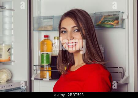 Frau trägt rotes Kleid öffnen Kühlschranktür und nach innen zu erreichen Blick auf Sie Kamera lächelnd. Aufnahmen in Innenräumen, horizontales Bild Stockfoto