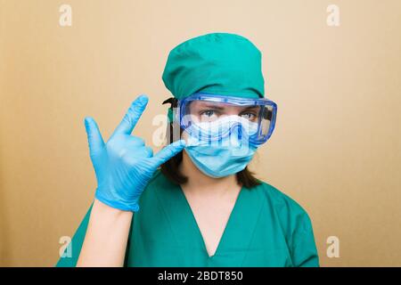 Junge Ärztin in schützenden Glasen grünen Anzug und Handschuhe fühlen sich glücklich, machen Rock oder Heavy Metal Zeichen mit der Hand. Stockfoto