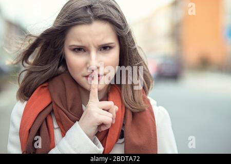 Shh Woman Wide Eyed bittet um Stille oder Geheimhaltung mit Finger auf Lippen Hush Hand Geste Stadtbild Outdoor-Hintergrund Hübsches Mädchen, das Finger auf li legt Stockfoto