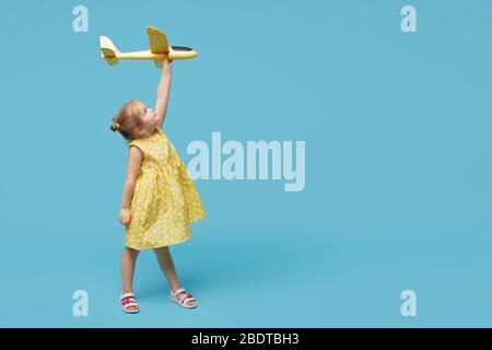 Kleines Mädchen Kind in einem Sommerkleid hält in seinen Händen ein Spielzeug Flugzeug auf einem blauen Hintergrund. Zeit für Reisen Stockfoto