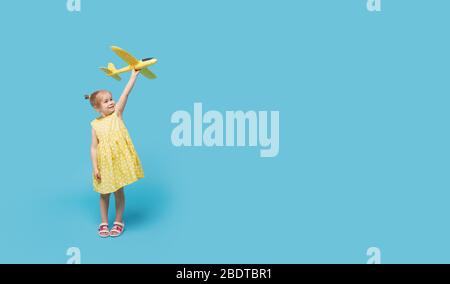 Kleines Mädchen Kind in einem Sommerkleid hält in seinen Händen ein Spielzeug Flugzeug auf einem blauen Hintergrund. Zeit für Reisen Stockfoto