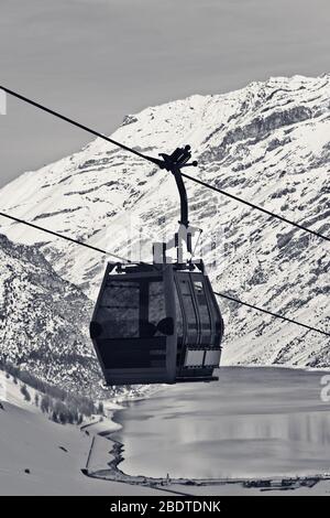 Gondelbahn, verschneite Piste abseits der Piste in hohen Winterbergen, Dorf und großer See im Hintergrund. Italienische Alpen am Abend. Livigno, Region Lomb Stockfoto