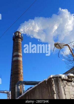 Rohre einer alten Fabrik werfen Wolken von giftigem weißem Rauch in den Himmel, die die Atmosphäre schädigen. Urbaner Smog aus Rauch aus Kesselhäusern. Weißer Rauch aus einem Kamin gegen einen blauen klaren Himmel. Stockfoto