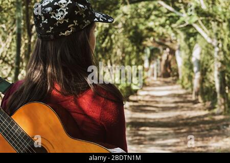 Junge kubanerin mit einer Akustikgitarre auf dem Rücken, die in eine Straße durch den Wald geht, die vor ihr liegt. Stockfoto