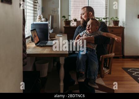 Vater arbeitet von zu Hause mit einem einjährigen Jungen weinend in seinem Schoß Stockfoto