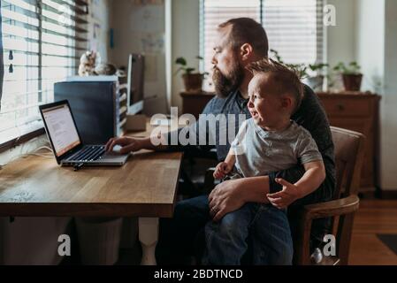 Vater arbeitet von zu Hause mit einem einjährigen Jungen weinend in seinem Schoß Stockfoto