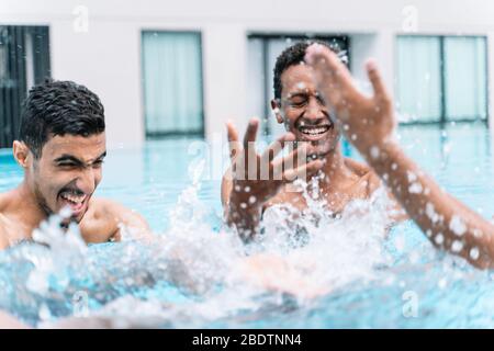 Junger Mann lächelnd, als er spielt und hebt die Hände in einem Kreis mit einer Gruppe von Freunden im Wasser eines Pools von Gebäuden umgeben Stockfoto