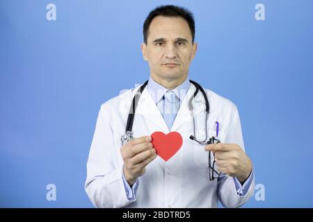 Arzt auf blauem Hintergrund hält Herz in den Händen zeigt es mit Zeigefinger Stockfoto