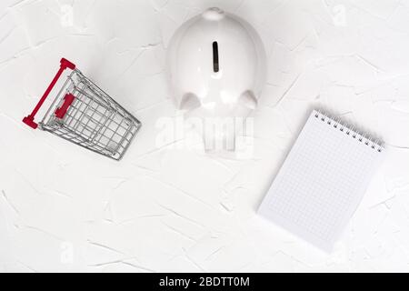 Weißes Sparschwein, Einkaufswagen und Notizbuch auf weißem Hintergrund. Blick von oben. Konzept speichern. Stockfoto