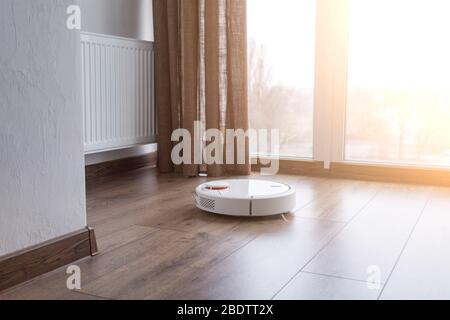 Weißer Roboter-Reiniger auf einem Holzboden in der Nähe des Panoramafensters. Haushaltsgeräte für die Reinigung des Hauses. Sonniger Tag. Stockfoto