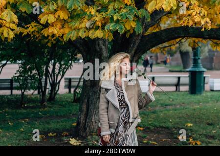 Junge blonde Mädchen zu Fuß in öffentlichen Park im Herbst und riecht rote Rose in der Hand Stockfoto