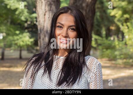Porträt von glücklichen indischen junge Frau lächelnd Blick auf Kamera mit dunklen langen Haaren isoliert grünen Bäumen Park Hintergrund Stockfoto