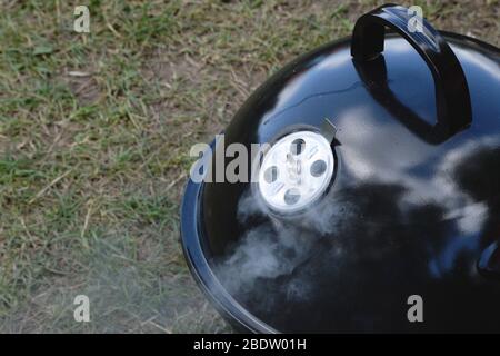 Kleine tragbare Holzkohle Grill auf dem Gras, mit Wolken reflektieren auf ihm. Kochen im Freien während des Campingausflugs oder des Tages im Park. Stockfoto