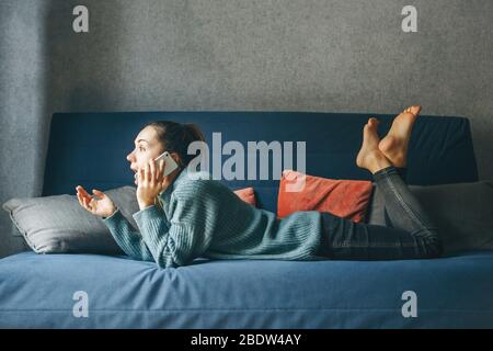 Mädchen, das auf einem Sofa liegt, benutzt ein Handy. Sie ist überrascht während des Gesprächs. Stockfoto