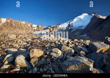 Wanderer in der gelben Jacke auf den Steinen vor hohen schneebedeckten Berge bei Mondaufgang Hintergrund Stockfoto