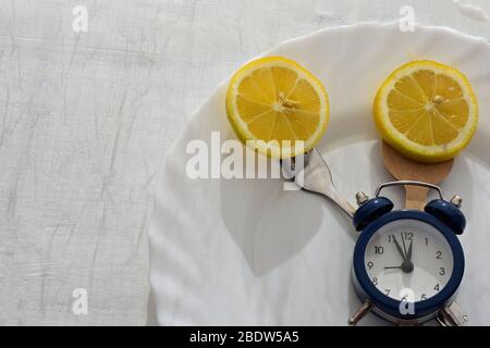 Frische Zitrone auf weißem Teller, Besteck und Wecker zum Abendessen oder Mittagessen/Zeit zum abnehmen/Essenskontrolle/Diätkonzept Stockfoto