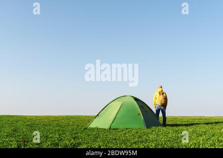 Tourist in gelben Jacke in der Nähe seines Zeltes auf Sommerfeld. Blauer Himmel. Reise- und Abenteuerkonzept Stockfoto