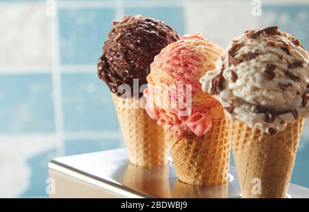 Drei köstliche Eiscreme-Aromen in Zapfen, die aufrecht in einem Metallhalter vor einem blau melierten Fliesenhintergrund mit Kopierraum in der Nähe dargestellt sind Stockfoto