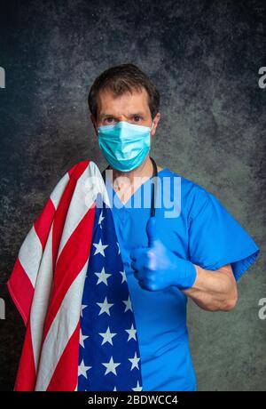 Doktor, der ein Daumen hoch Handzeichen tut, das blaue Krankenhaus-Peelings mit Gesichtsmaske und Stethoskop trägt, die amerikanische Flagge nahe an seiner Brust haltend. Stockfoto