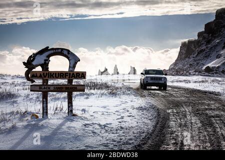 Ein Auto fährt am Schild vorbei zum schwarzen Sandstrand Vikurfiara mit Reynisdrangar Basaltsteinhaufen in der Ferne bei Vík í Mýrdal in Island Stockfoto